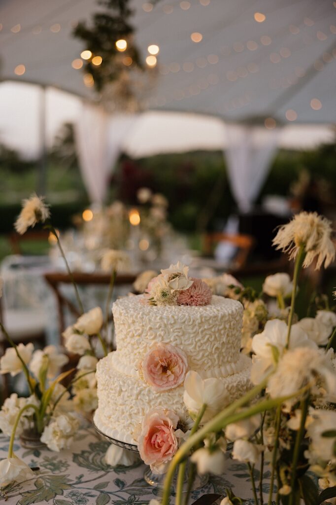 A wedding cake sits on a table under a tent surrounded by white flowers 