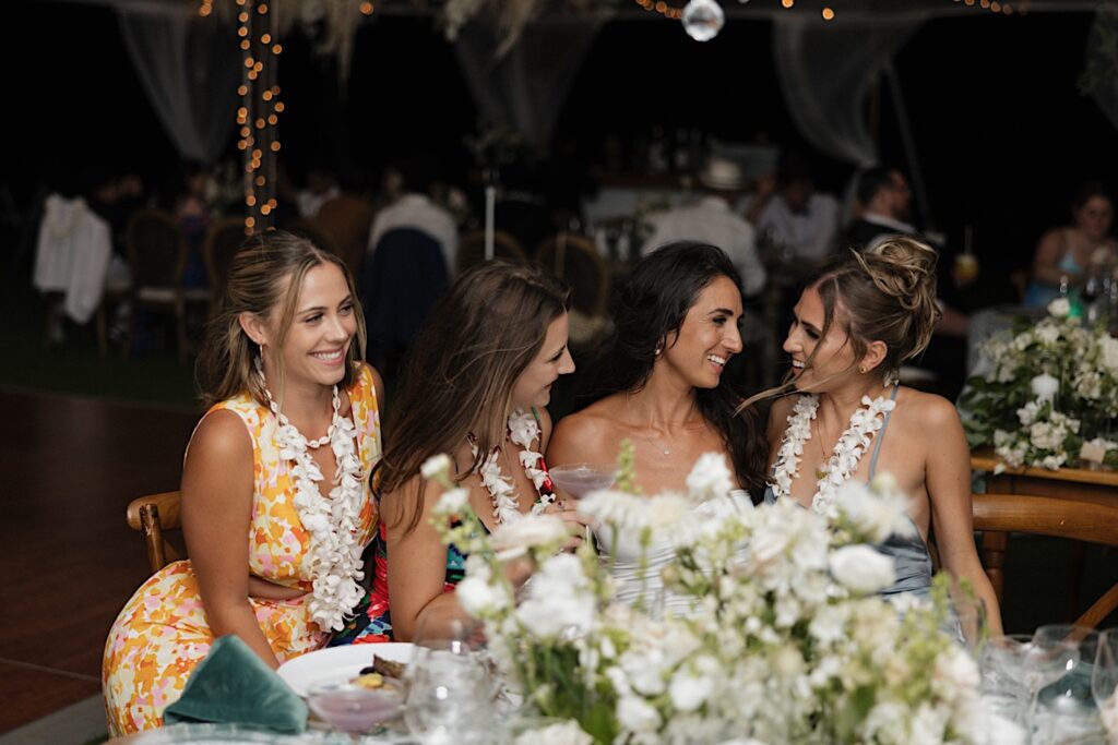 A bride sits with 3 members of her wedding at a table during the reception and laugh with one another under a tent at the venue Kukui’ula on Kauai