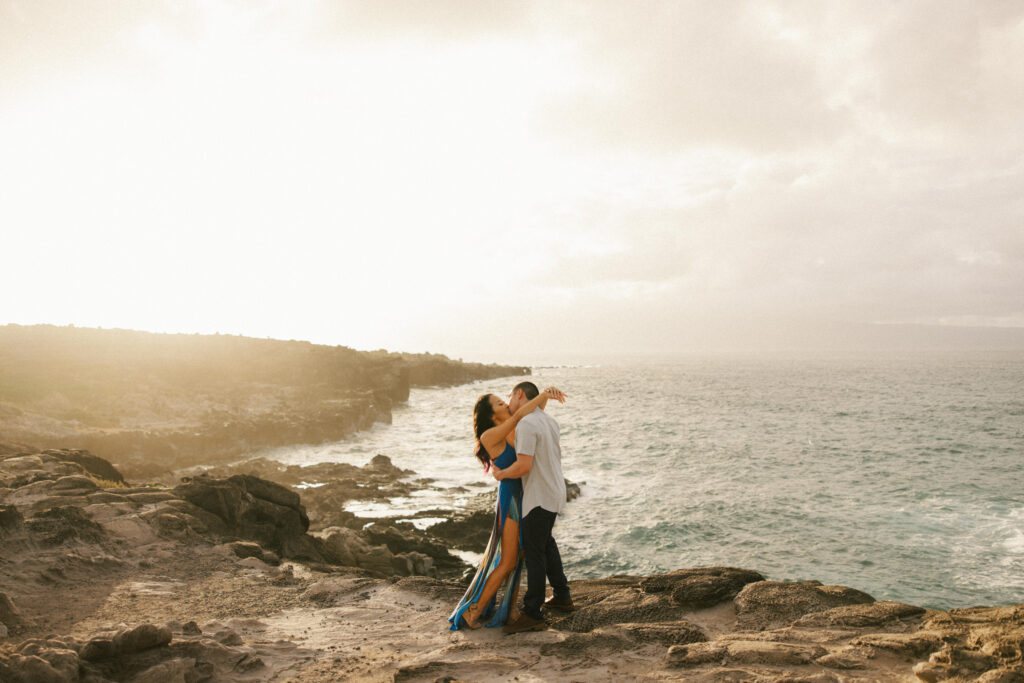 During their engagement session on the island of Maui a woman smiles up at a man as she lays her arms around his neck, with the island of maui in the distance.