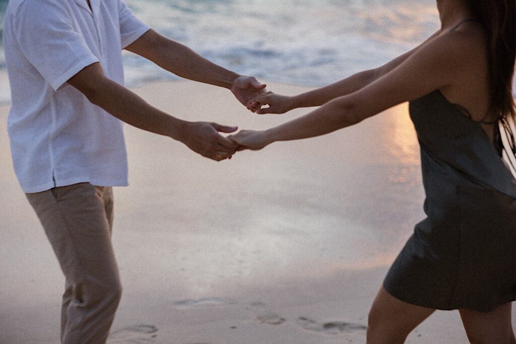 A man and woman hold hands while on a beach of Oahu as the sun sets while they take their engagement photos