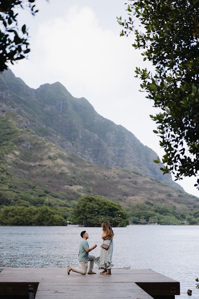 A man gets down on one knee to propose to a woman while they stand on a dock looking out over a lake and mountains in the background