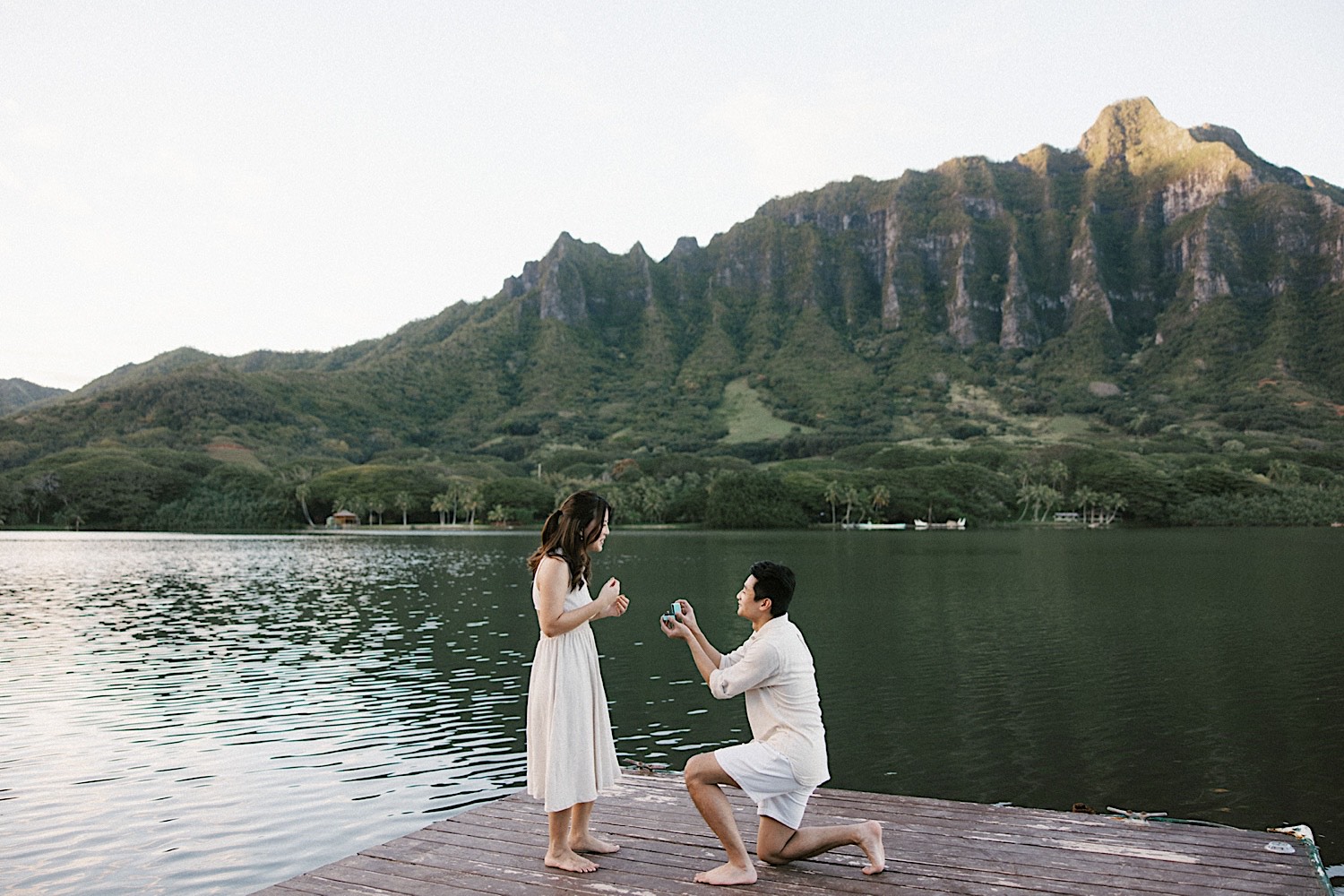 A woman smiles as a man gets down on one knee in front of her for their proposal at secret lake on Oahu