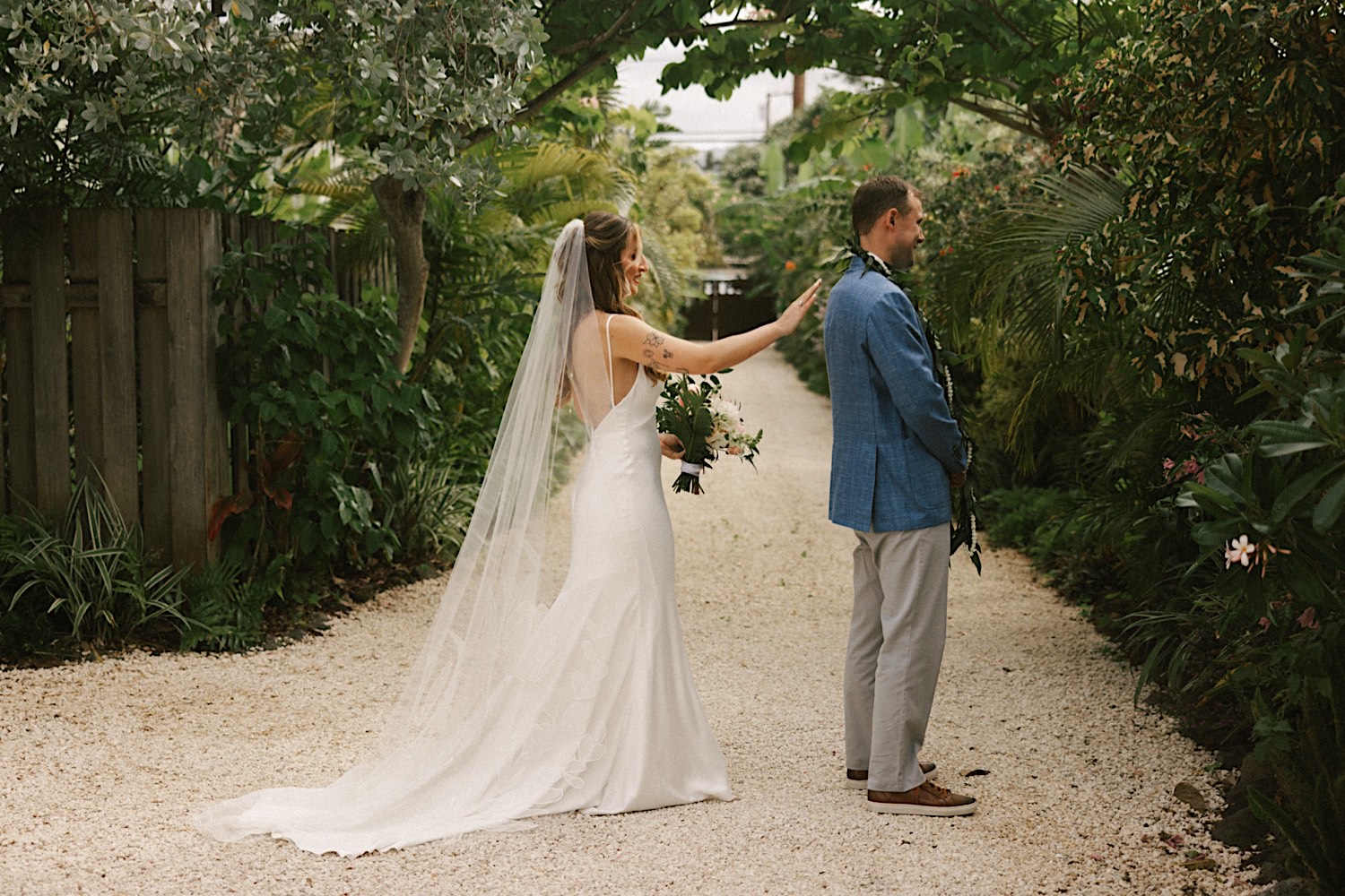 A bride is about to tap the groom on his shoulder for their first look on their wedding day in Hawaii, they couple is standing on a stone path surrounded by greenery