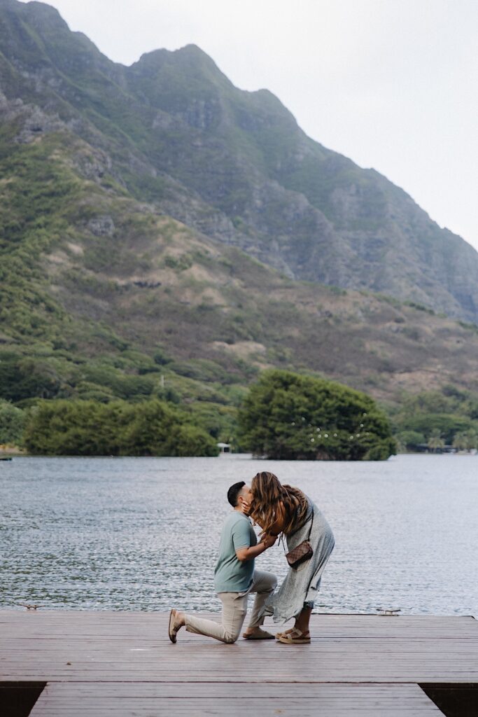 A woman leans over to kiss a man who's down on one knee after proposing to her on a dock looking out over a lake and mountains