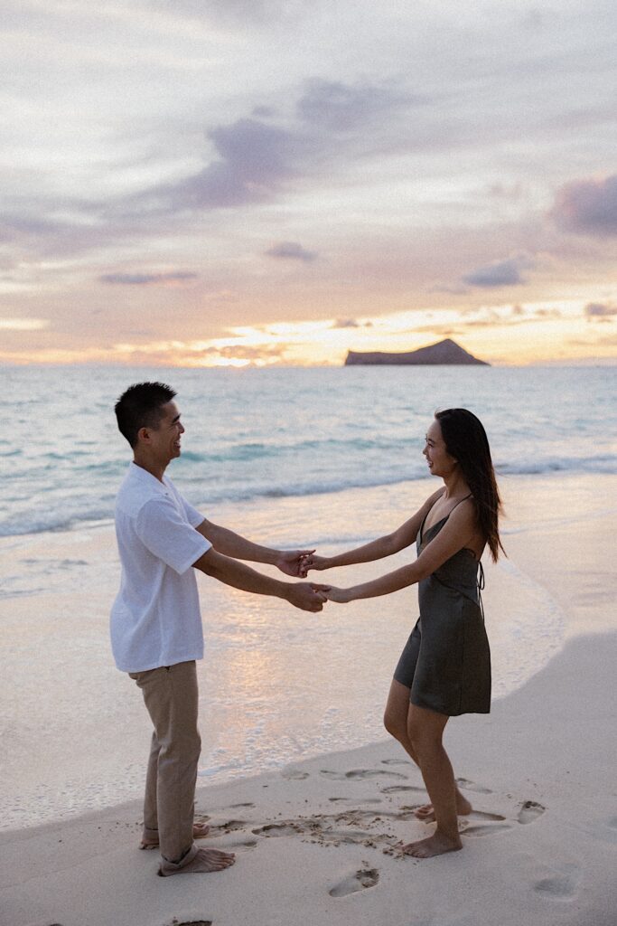 A man and woman on a beach of Oahu hold hands and smile at one another as the sun sets behind them