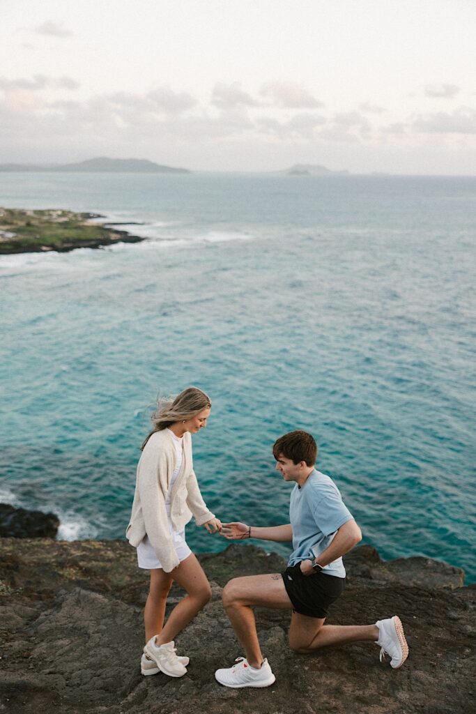 A man down on one knee holds a woman's hand as he reaches for an engagement ring in his pocket, the couple is on top of a cliffside in Oahu looking out over the ocean