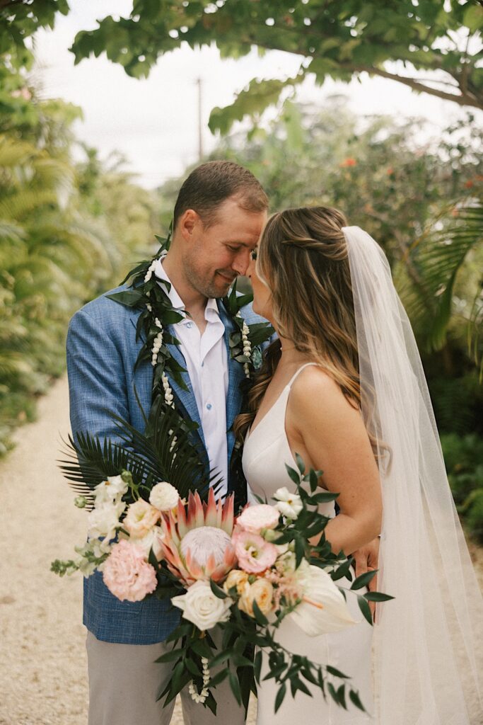 A bride and groom smile at one another while touching noses on a path surrounded by greenery