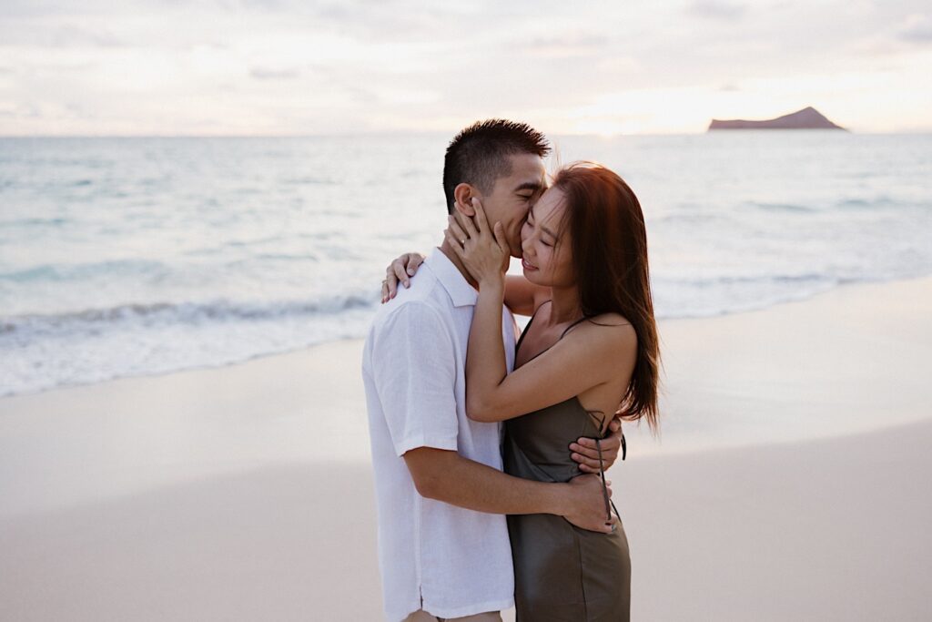 A man kisses a woman on the cheek while they hug on a beach in Oahu at sunset during their engagement photos session