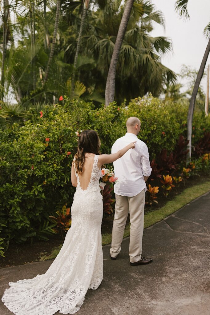 A bride touches a groom on his shoulder to get him to turn around for their first look while standing on a sidewalk surrounded by lush greenery