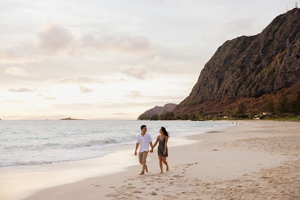 While taking engagement photos a man and a woman hold hands while walking along the beach of Oahu at sunset