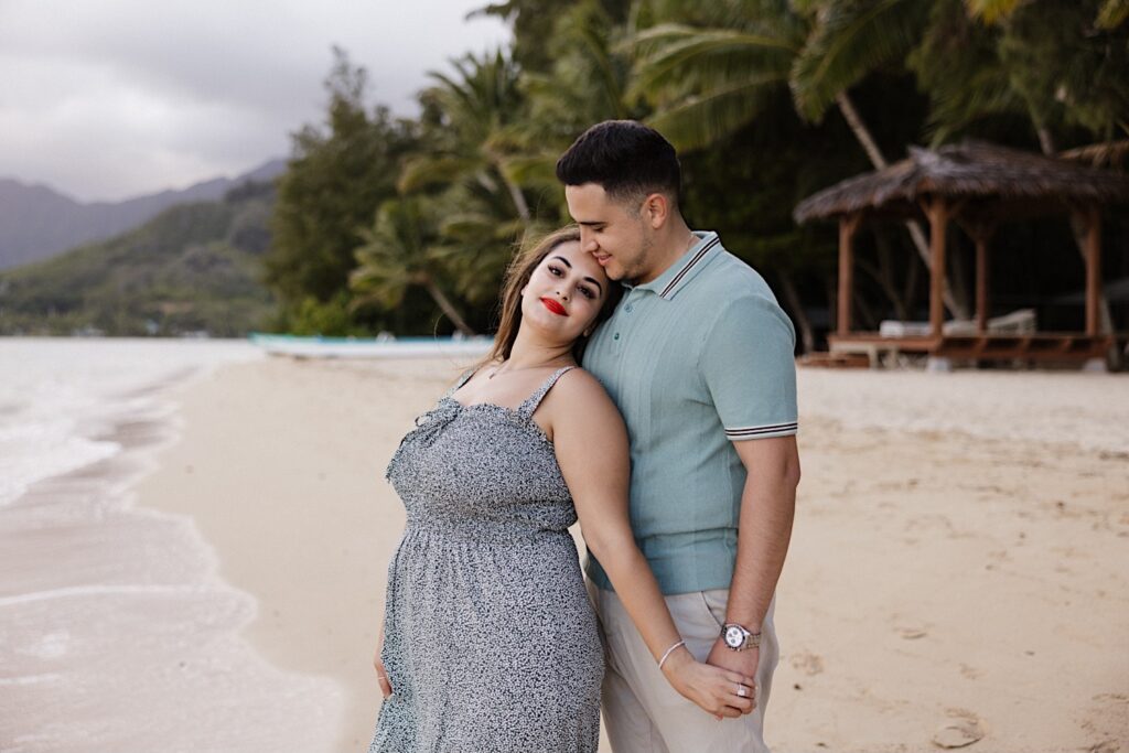 After their proposal at Secret Island on Oahu a man and woman stand on a beach together, the woman looks at the camera while standing in front of the man who looks down towards her