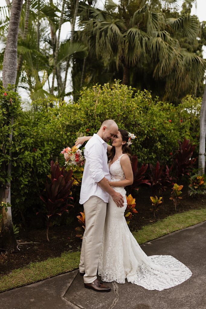 A bride and groom embrace and smile at one another while standing on a sidewalk with flowers and greenery behind them