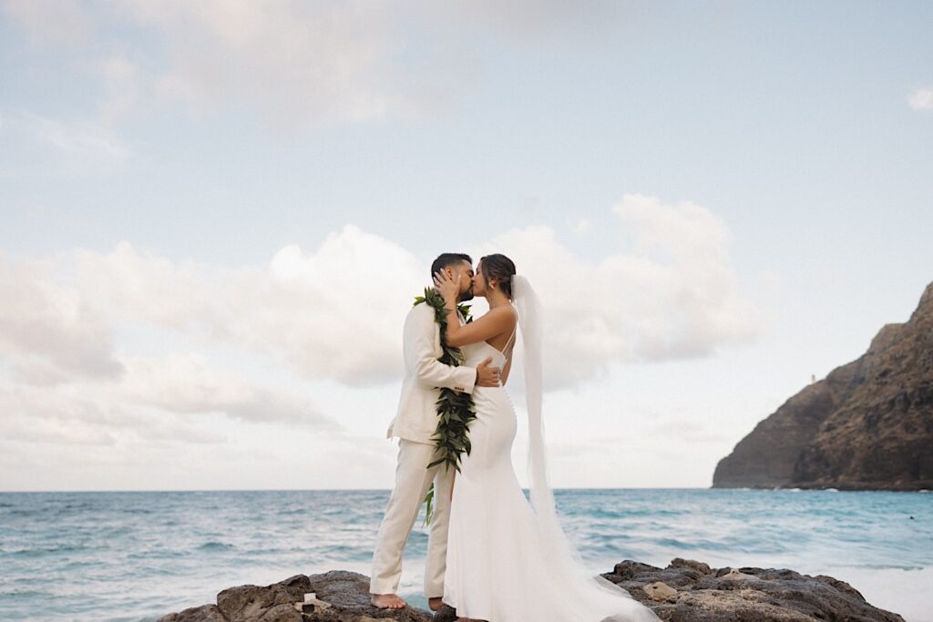 A bride and groom kiss one another after their first look on their wedding day while standing on a rock formation on the ocean shore of Hawaii