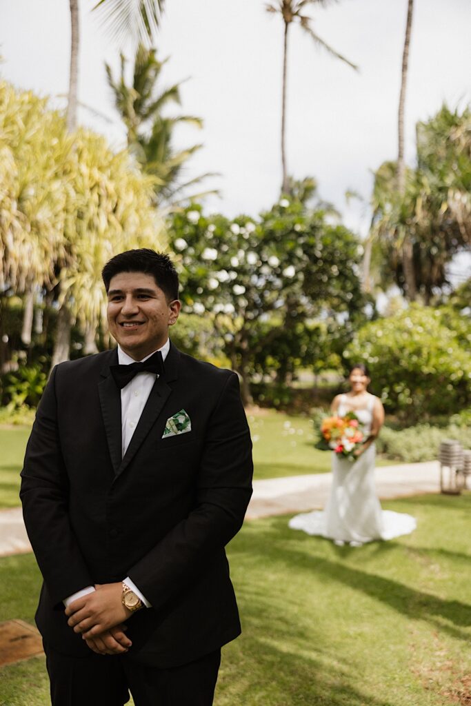 A groom smiles while the bride stands behind him as she is about to approach him for their first look on their wedding day in Hawaii