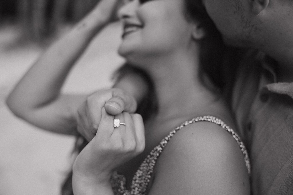 Black and white photo of a woman smiling as a man hugs her from behind while she shows her new engagement ring from their proposal on Oahu to the camera