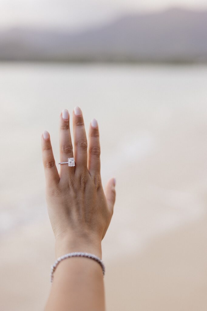 A woman's hand shows off an engagement ring with the ocean in the background