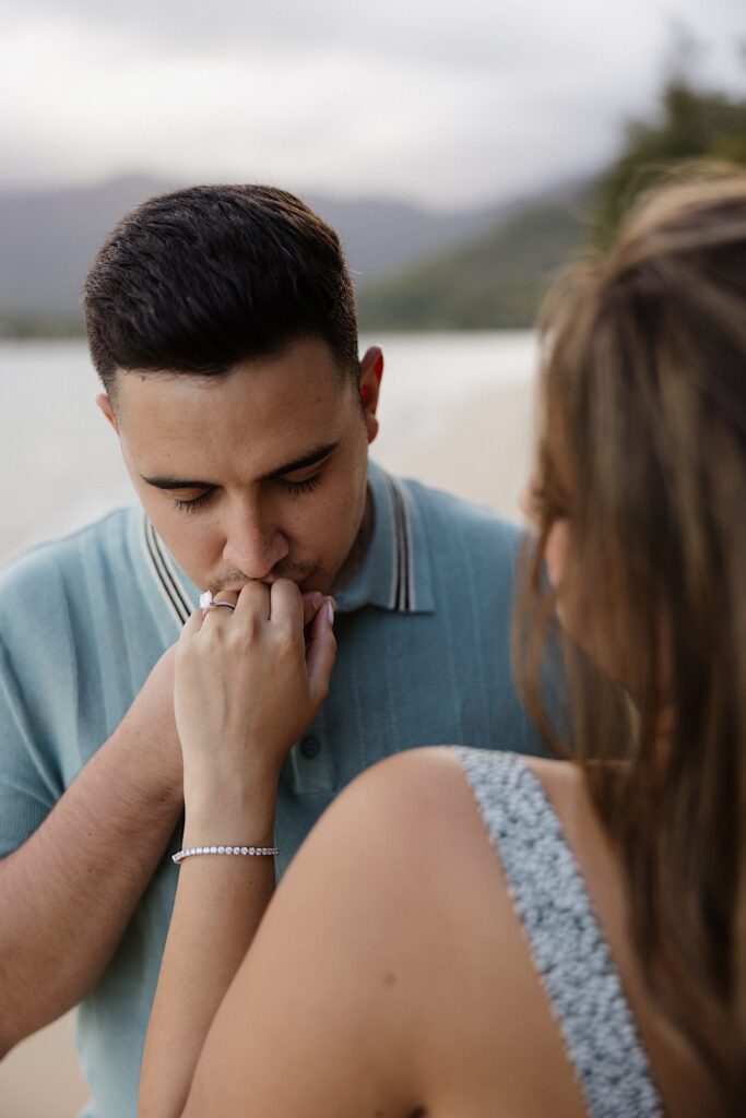 A man kisses a woman's hand with an engagement ring on it as the woman has her back to the camera