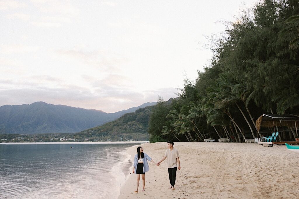 A couple hold hands and look at one another while walking along a beach on Oahu next to the ocean after their proposal