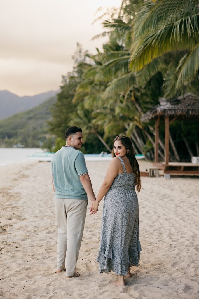 A man and woman hold hands and walk on a beach together away from the camera, the man is smiling at the woman while she smiles back at the camera