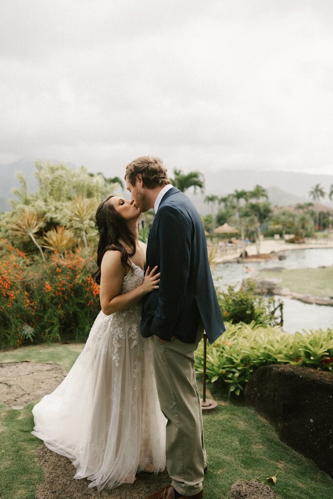 A bride and groom kiss on a cloudy day surrounded by tropical vegitation