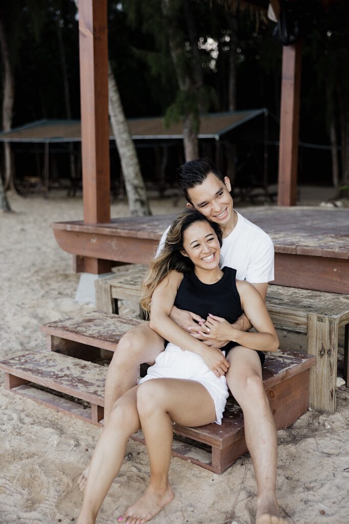 A man and woman sit together on wooden steps leading to a beach, they smile as the man hugs the woman from behind while she sits between his legs
