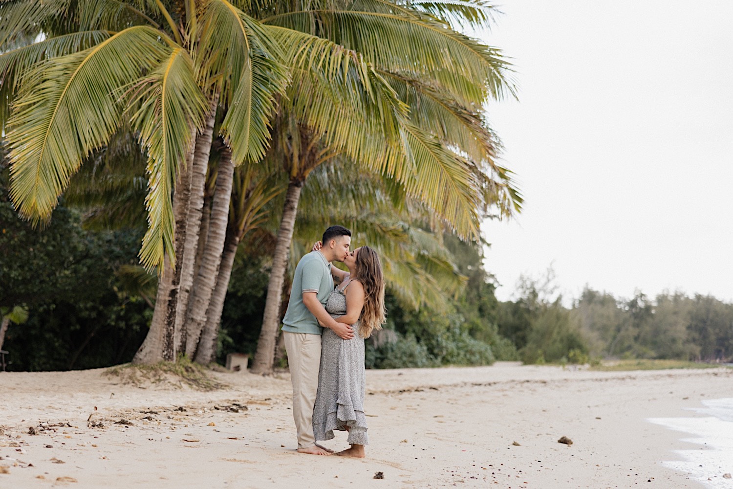 After their proposal at Secret Island on Oahu a man and woman kiss one another while standing under a palm tree on the beach