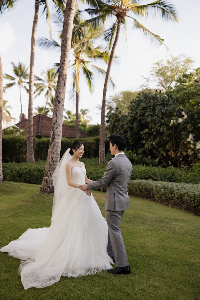 A bride and groom hold hands and smile at one another while standing in a grassy field surrounded by palm trees