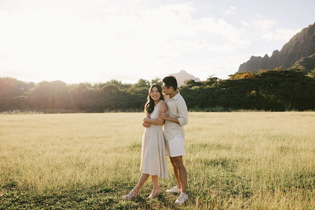 A woman smiles towards the camera while a man hugs her from behind and looks at her, they're standing in a field with the mountains of Oahu behind them and the photo was taken after their proposal