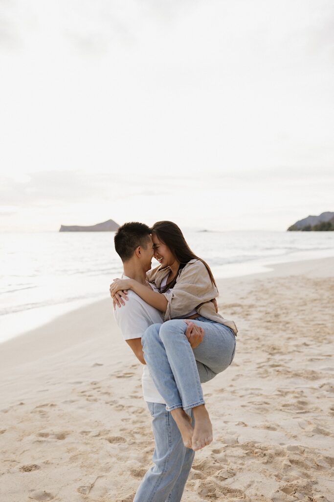A man hold a woman in his arms as they smile at one another while on the beach of Oahu