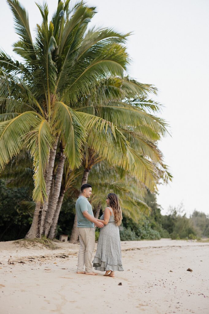 A man and woman hold hands and smile at one another while standing underneath a palm tree on the beach