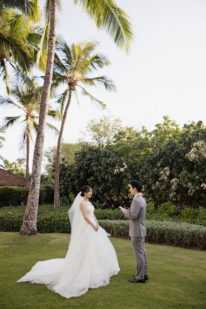 A bride smiles as the groom reads his vows to her while they stand in a grass clearing surrounded by palm trees