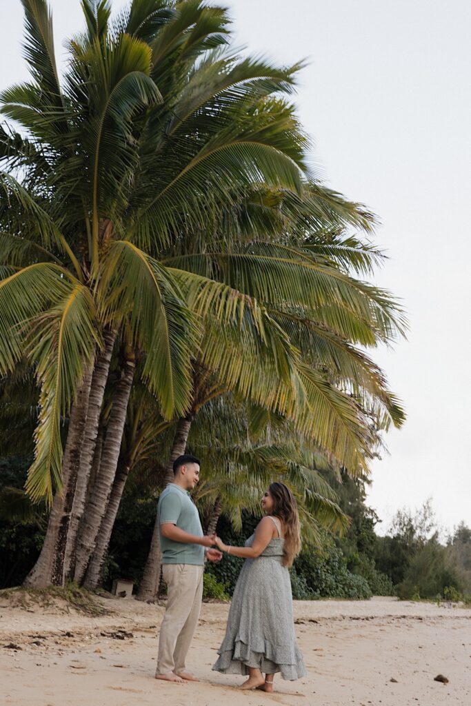 A man and woman hold hands while standing on a beach together underneath a palm tree