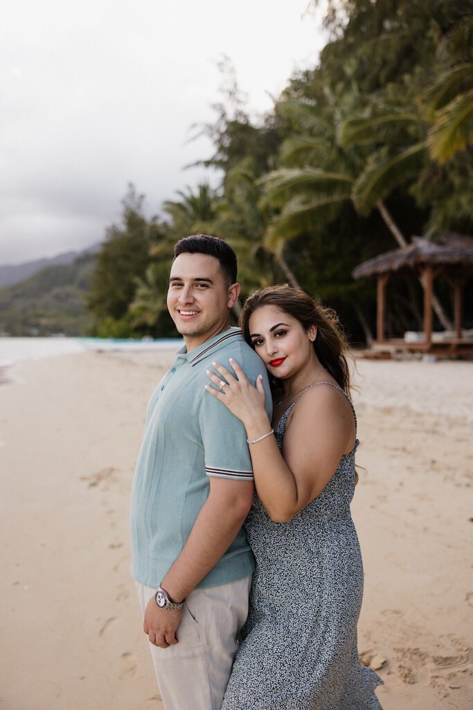 A woman hugs a man from behind while they both smile at the camera while standing on a beach