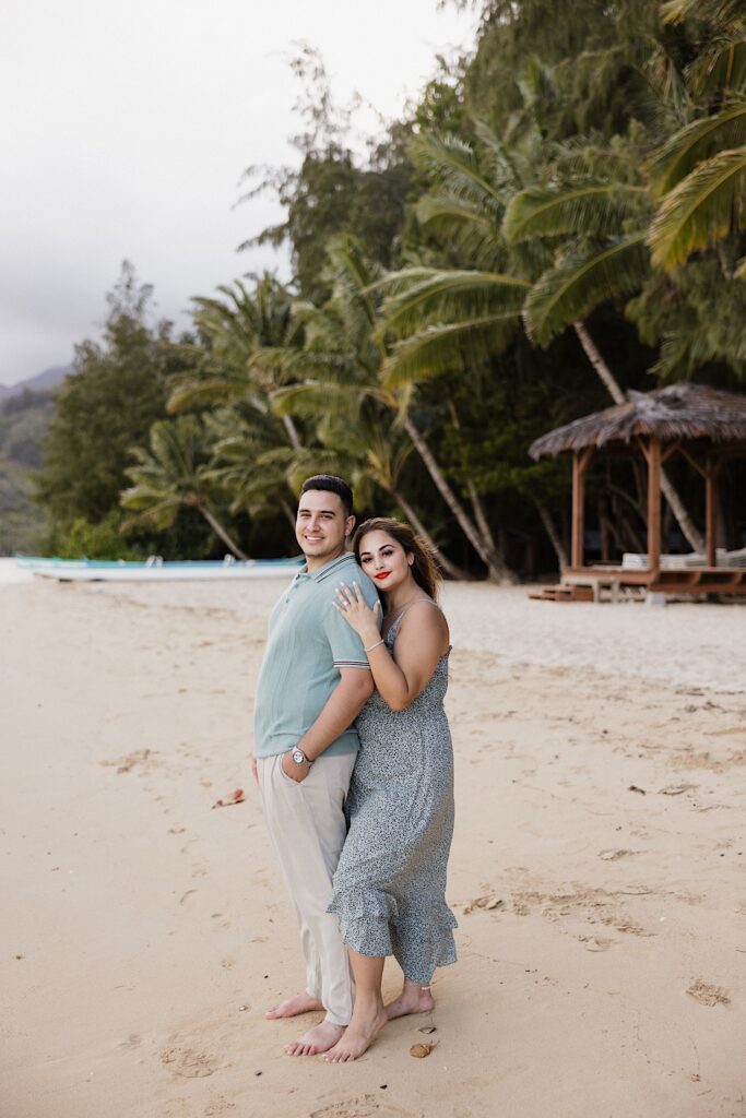 A woman hugs a man from behind while they both smile at the camera while standing on a beach