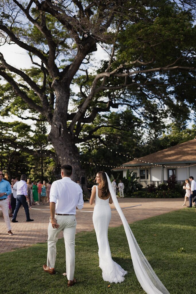 A bride and groom walk together towards their outdoor wedding reception under a massive tree with string lights attached to it