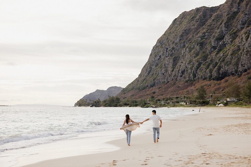 A man and woman walk hand in hand away from the camera along a beach of Oahu during their engagement photos 