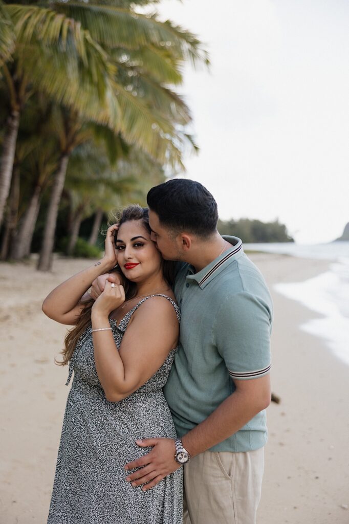A woman smiles at the camera while a man hugs her from behind and kisses her cheek as the two stand on a beach together