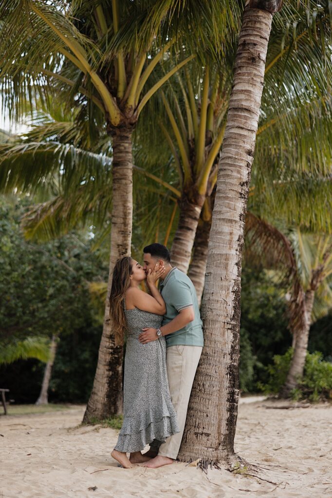 A man leans against a palm tree on a beach as a woman kisses him and he holds her 