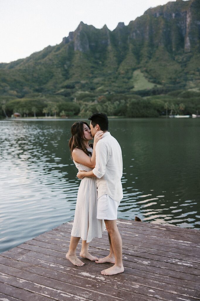 A man and woman kiss one another while on a dock of a lake with mountains in the background