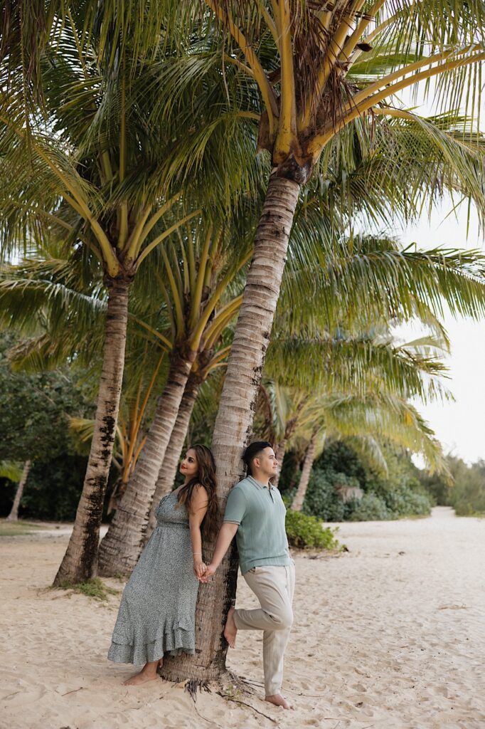 A man and woman stand against a palm tree on the beach and hold hands while looking in opposite directions