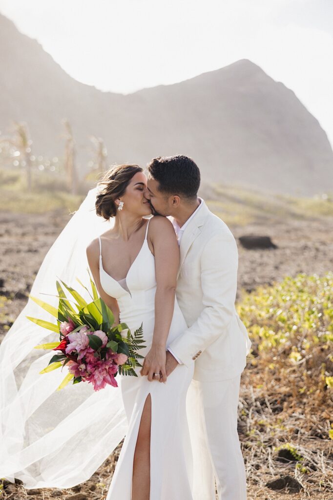 A groom kisses the bride while standing behind her as they stand in a clearing in front of a mountain
