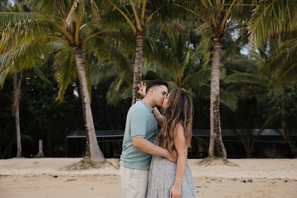 After their proposal at Secret Island on Oahu a man and woman embrace and kiss one another while on a beach with palm trees behind them