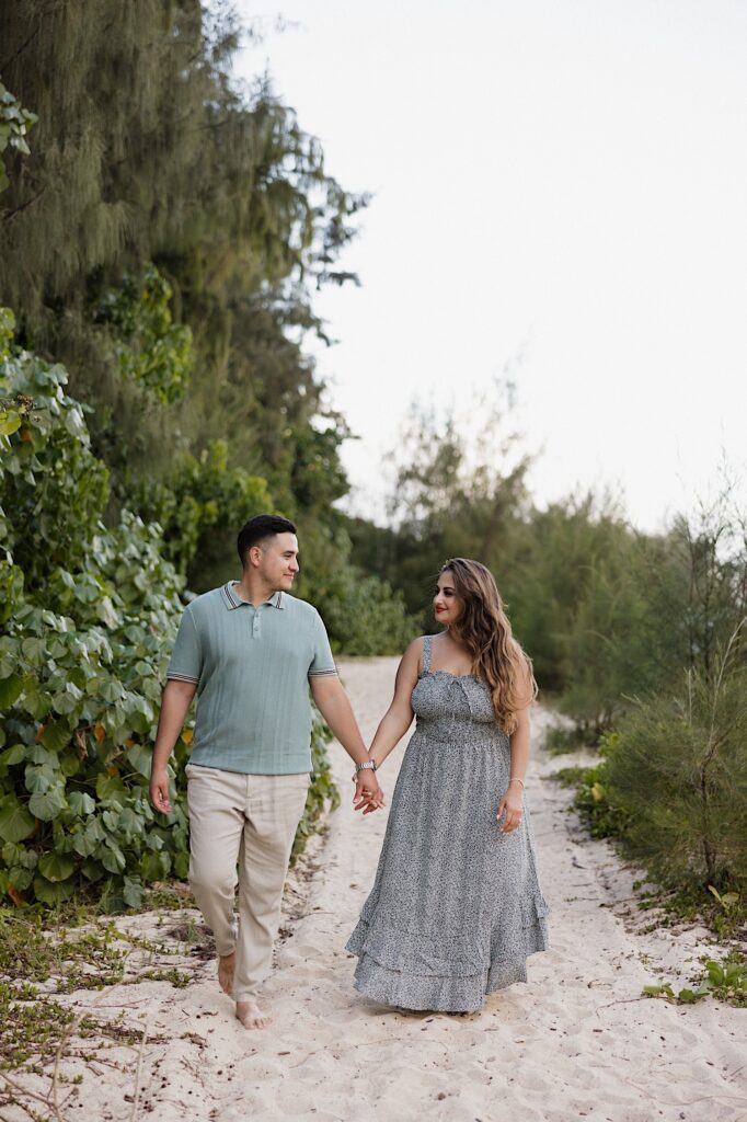 A man and woman walk hand in hand towards the camera while smiling at each other, they're on a sandy path next to some trees and other vegetation