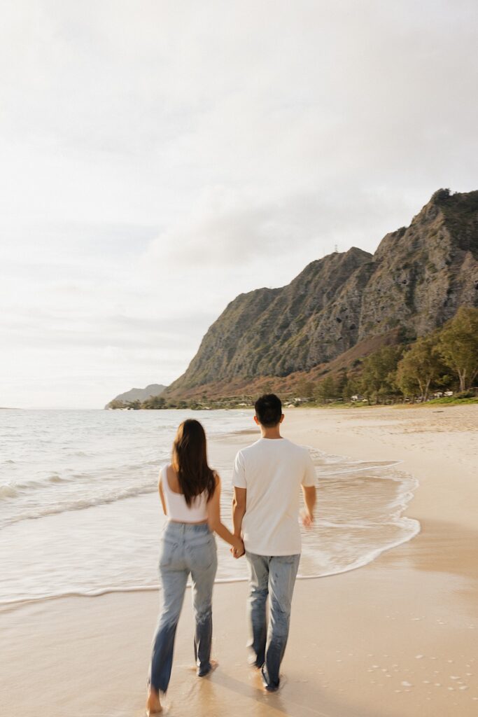 A couple walk hand in hand along a beach of Oahu towards mountains in the background at sunset