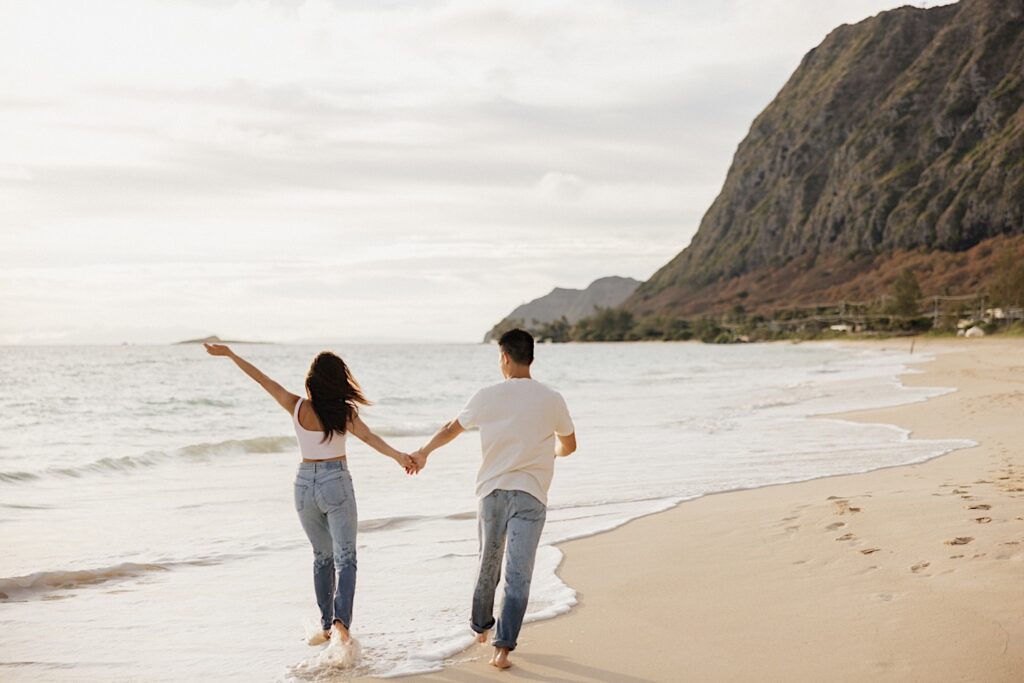 While taking engagement photos on Oahu a man and woman hold hands and walk along the beach together