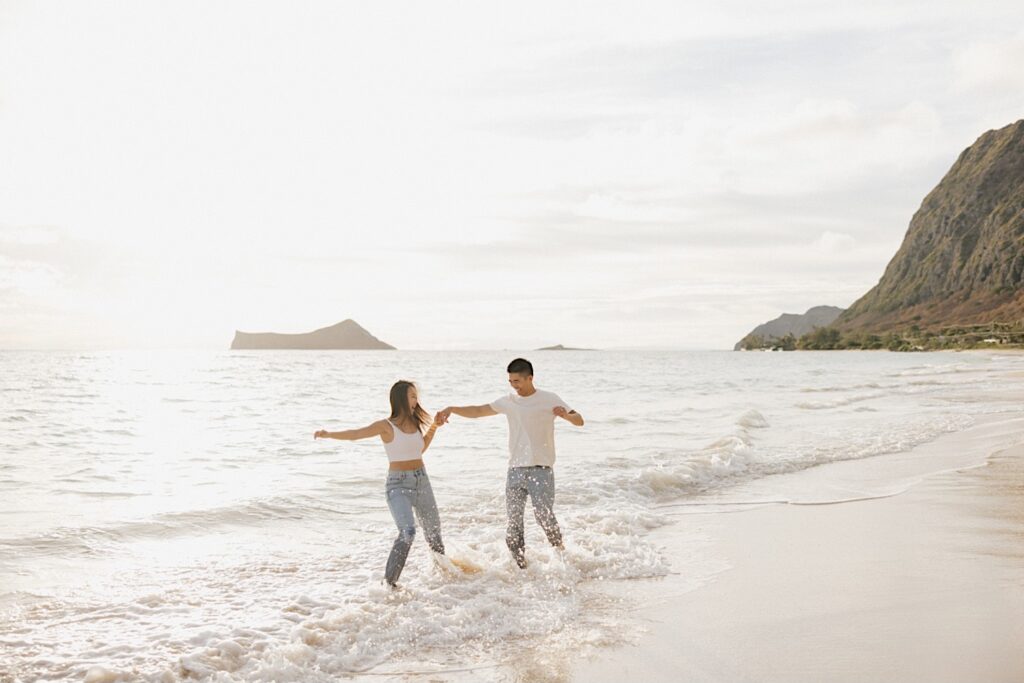 A man and woman dance together in the water of the ocean while on a beach of Oahu during their engagement photos session