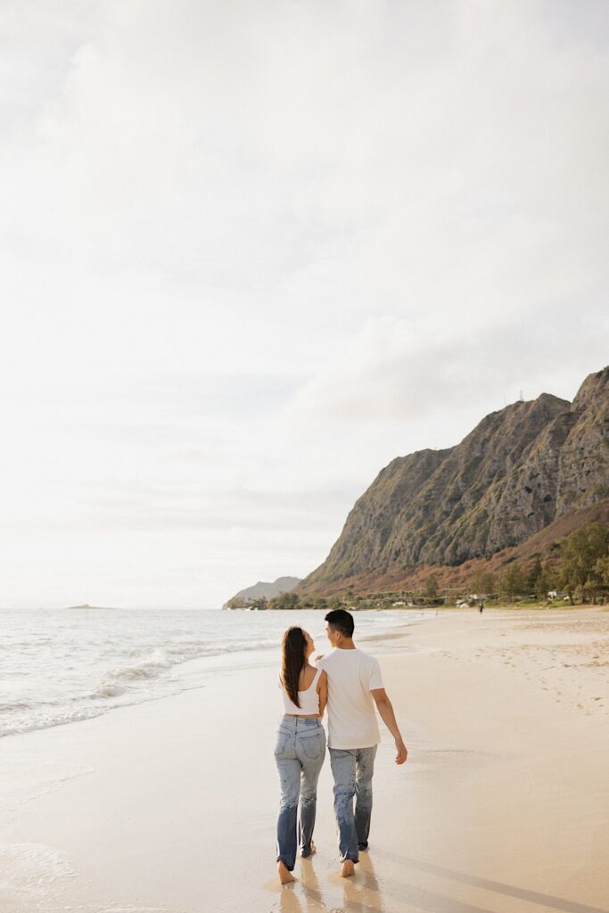 A man and woman walk along a beach side by side at sunset towards mountains in the distance on the islands of Oahu