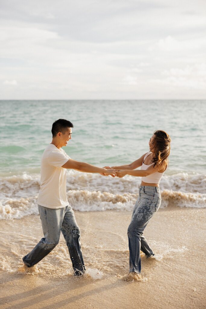 A man and a woman are holding hands and dancing together on the beach as the water splashes around them at sunset