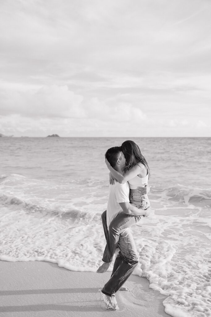 Black and white photo of a man holding a woman in the air as they're about to kiss while on a beach of Oahu