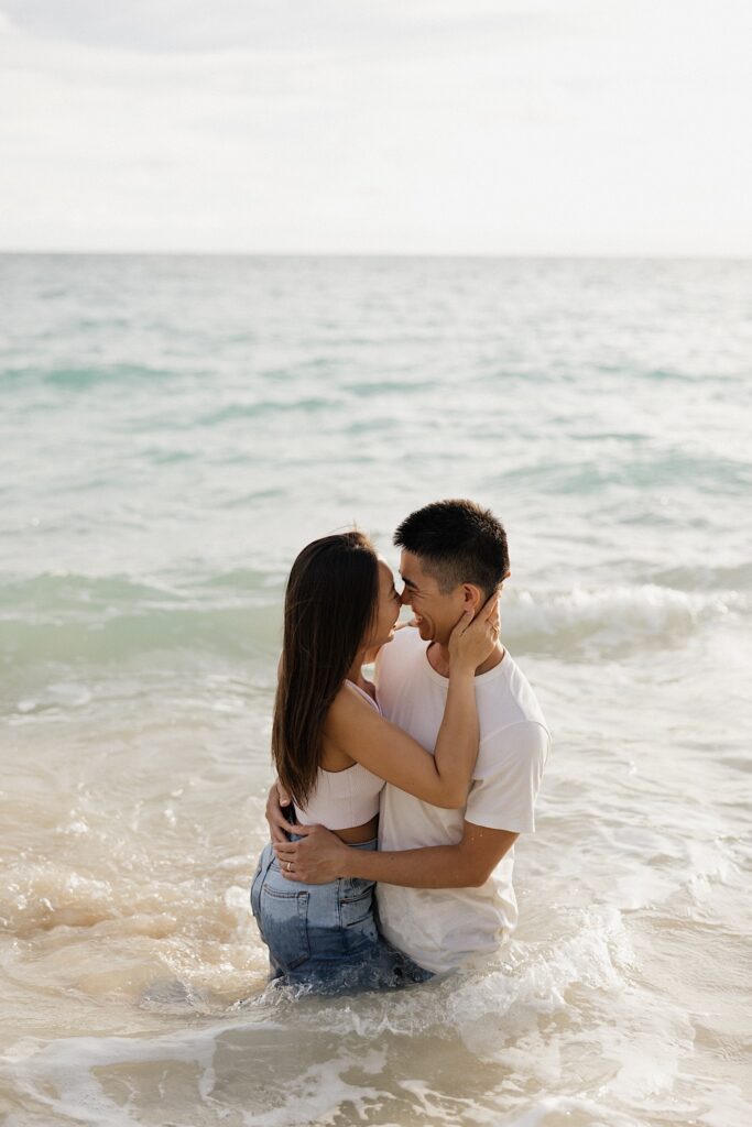 A man and woman smile and embrace while about to kiss one another as they kneel in the ocean on a beach of Oahu